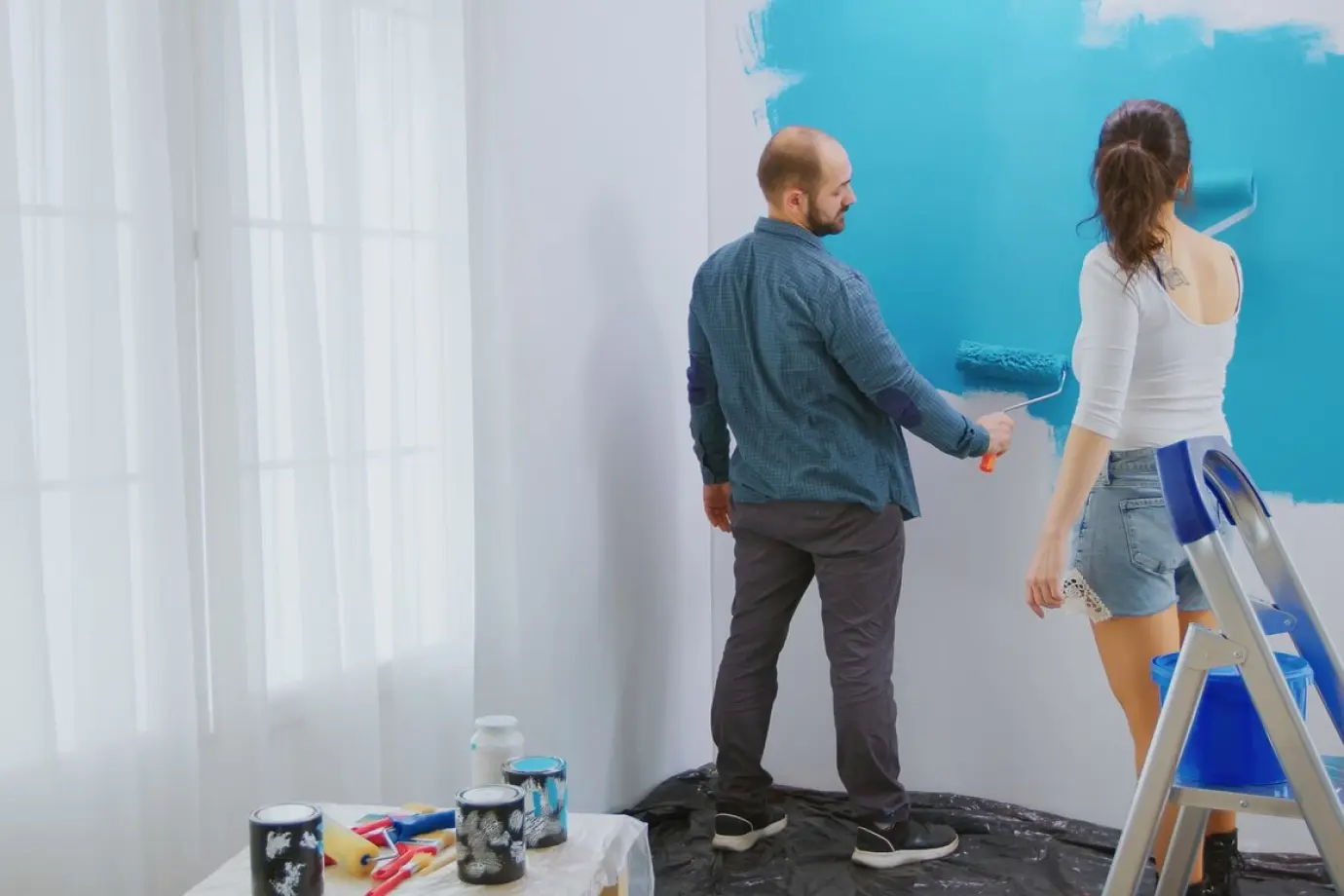 A-couple-painting-a-wall-blue-man-holds-roller-up-high-woman-reaches-for-roller-both-standing-on-plastic-covered-floor