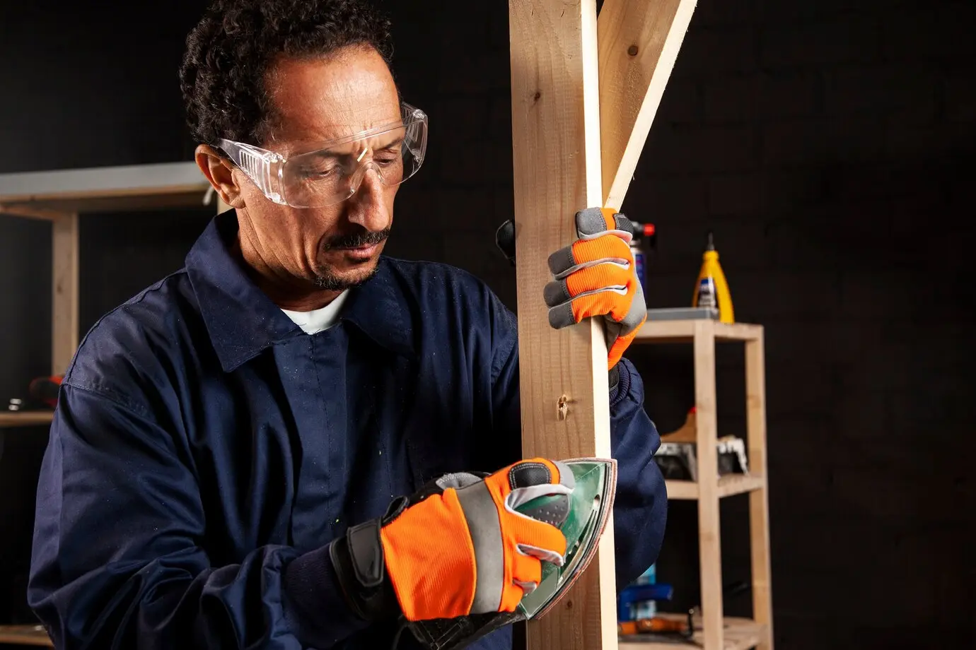 A-handyman-in-safety-goggles-and-orange-gloves-carefully-sands-a-wooden-plank-in-his-workshop
